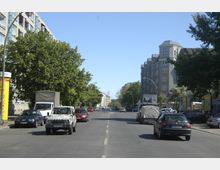 Straßenszene in einer Stadt mit mehrspuriger Fahrbahn, flankiert von Wohn- und Bürogebäuden; links moderne Häuser mit großen Fenstern und rechts ein graues, mehrstöckiges Gebäude mit Mansardendach. Parkende Autos und Bäume säumen beide Straßenseiten, bei klarem Himmel und Tageslicht.