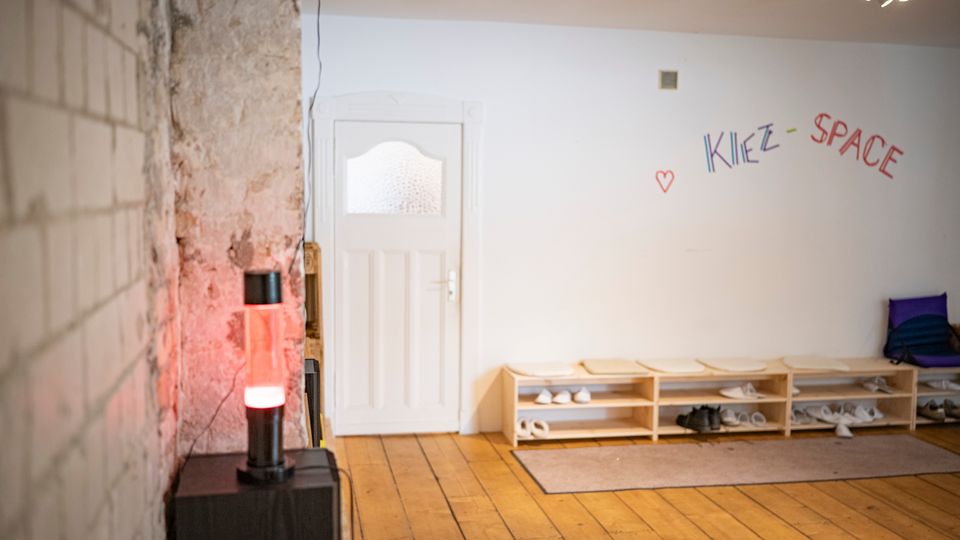 Eingangsbereich eines Gemeinschaftsraums mit Holzdielenboden, weißer Tür und einem niedrigen Schuhregal entlang der Wand. An der Wand steht „KIEZ-SPACE“ in bunten Buchstaben; links sind freigelegte, raue Mauerflächen und eine leuchtende Lampe zu sehen.