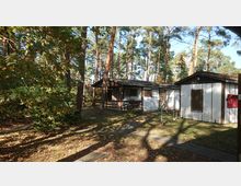 Ein Holzbungalow mit überdachter Terrasse steht in einem lichten Nadelwald. Umgeben von Bäumen und Laub befindet sich daneben ein kleiner Schuppen; im Vordergrund sind Gehwegplatten und ein Wasseranschluss zu sehen.