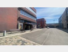 Straßenszene mit dem Eingang zur U-Bahn-Station „U-Bhf Klosterstraße“ in einem modernen, rotbraunen Gebäude auf der linken Seite. Im Hintergrund sind gemischte Architektur, ein rotes historisches Gebäude und eine Eisenbahnbrücke mit Graffiti zu sehen, daneben steht ein Baukran.