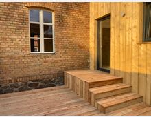 Backsteinfassade eines Gebäudes mit einem modernen Holzanbau. Im Vordergrund eine Terrasse aus Holz, mit einer kurzen Treppe, die zu einer Glastür führt; daneben ein rundbogiges Fenster mit weißem Rahmen.