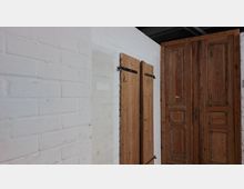 Weiße Ziegelwand mit zwei Holzöffnungen: Links eine doppelflügelige Holztür mit schwarzen Scharnieren, rechts eine größere zweiflügelige Holztür mit dekorativen Paneelen. Oberhalb der Türen sind dunkle Rohre zu sehen.