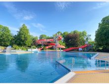 Ein Freibad mit einem großen Schwimmbecken, umgeben von Bäumen. Im Hintergrund befinden sich rote Wasserrutschen und Wasserspiele, wie ein Pilzbrunnen und kleinere Wasserfälle.
