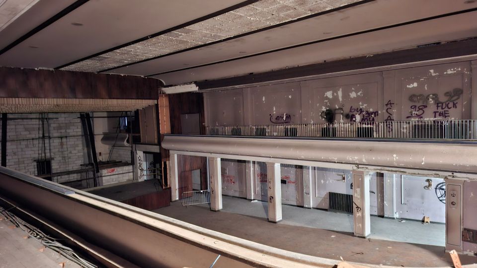 Blick in eine verlassene, stark heruntergekommene Innenhalle mit zwei Ebenen, umlaufender Galerie und Geländern. Abblätternde Wandverkleidungen, Graffiti, Säulenreihen und eine ehemalige Bühne oder Schalterzone sind sichtbar.