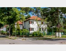 Ecke eines Wohnviertels mit einem zweistöckigen weißen Einfamilienhaus mit roten Dachziegeln, umgeben von Bäumen und einem grünen Zaun. Im Vordergrund sind Straßenlaternen, ein Gehweg und ein Straßenschild sichtbar.