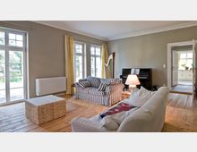 Wohnzimmer mit Holzboden und großen Fenstern, die viel Tageslicht hereinlassen. Der Raum ist möbliert mit einer weißen Couch, einem gestreiften Sofa, einem geflochtenen Couchtisch, einer Harfe und einem schwarzen Klavier.