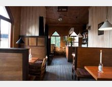 Ein rustikales Café oder Restaurant mit Holzwänden und dunklen Holzmöbeln. Die Einrichtung umfasst Sitznischen, einfache Holztische, weiße Hängelampen und Kerzen auf den Tischen, mit einem Fenster im Hintergrund, das Blick nach draußen bietet.