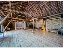 Geräumiger Innenraum einer rustikalen Scheune mit sichtbaren Holzbalken, einem hohen Dachgebälk und hellen Holzdielen. Im Hintergrund sind dekorative Lichterketten, Möbel aus Holz und Korb sowie vintage-inspirierte Sitzgelegenheiten sichtbar.