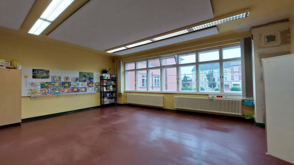Klassenzimmer mit gelb gestrichenen Wänden, großen Fenstern auf der rechten Seite, die den Blick auf ein Gebäude und Bäume freigeben. An der linken Wand befindet sich eine Pinnwand mit Kinderzeichnungen sowie ein Regal mit Büromaterialien, und der Boden ist rötlich-braun.