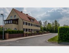Zweistöckiges Wohnhaus in hellgelber Farbe mit Satteldach und mehreren Fenstern mit braunen Fensterläden, umgeben von einem Garten und einem niedrigen Zaun. Rechts führt eine asphaltierte Straße vorbei, im Hintergrund befindet sich eine Baumgruppe unter bewölktem Himmel.
