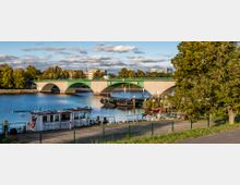 Brücke über einen Fluss mit mehreren Bögen, im Vordergrund ein Uferbereich mit festgemachten Booten und einem Zaun, im Hintergrund Gebäude und Bäume. Der Himmel ist bewölkt mit vereinzeltem Sonnenschein.