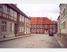Straße in einer Altstadt mit Kopfsteinpflaster, flankiert von historischen Fachwerk- und Putzgebäuden. Im Hintergrund steht ein großes Fachwerkhaus mit rotem Ziegeldach und zahlreichen weißen Fenstern.