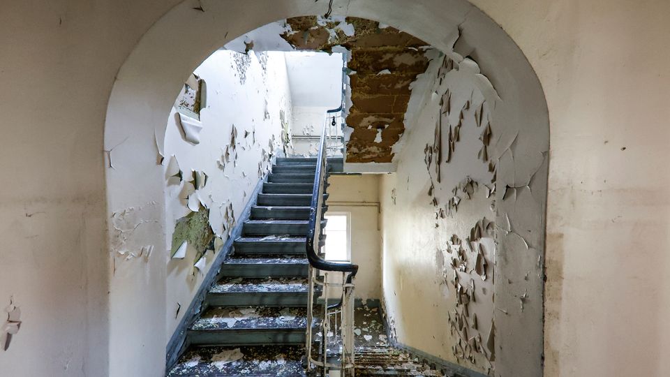Blick in ein verfallenes Treppenhaus mit grauen Stufen und Geländer unter einem Rundbogen. An den Wänden und der Decke blättert großflächig die Farbe ab, und am oberen Ende fällt Licht durch ein Fenster.