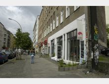 Straßenszene in einer urbanen Umgebung mit mehrstöckigen Wohngebäuden auf der rechten und linken Seite. Im Vordergrund befinden sich ein Gehweg, geparkte Autos, Bäume und ein Geschäft mit großen Fenstern und einer Werbetafel.