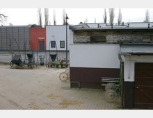 Ein gepflasterter Hof mit Karren und Schubkarren, umgeben von modernen und älteren Gebäuden, darunter eine teils verputzte Mauer mit sichtbarem Mauerwerk. Im Hintergrund stehen schlanke Bäume und ein modernes Gebäude mit roten und weißen Fassaden.