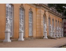 Gelb verputztes historisches Gebäude mit hohen, verzierten Fenstern und einer Reihe von weißen Marmorskulpturen auf Sockeln entlang der Fassade; die Umgebung besteht aus einem Kiesweg und vereinzeltem Grün.