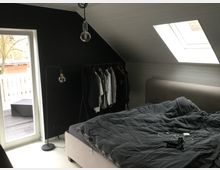 Schlafzimmer mit schräger Decke und Dachfenster, schwarzer Wand, einem Doppelbett mit grauer Bettwäsche und freistehendem Kleiderständer. Links führt eine Glastür mit Ausblick auf einen Balkon mit weißen Geländern.