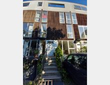 Moderne Wohnhausfassade mit vertikalen Holzlamellen und großen Glasfenstern. Vor dem Eingang mit Treppenstufen befindet sich ein kleiner, begrünter Bereich; ein Auto ist rechts geparkt.