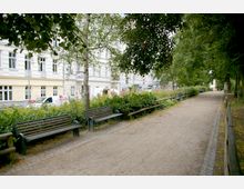 Ein kleiner Parkweg mit Bänken auf beiden Seiten, umgeben von Bäumen und Sträuchern. Im Hintergrund sind mehrstöckige Altbaugebäude mit weißen Fassaden und einige parkende Autos zu sehen.
