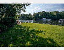 Rasenfläche am Ufer eines Flusses, umgeben von Bäumen. Im Hintergrund sind Wasserfahrzeuge und eine von Bäumen gesäumte Uferlinie zu sehen.