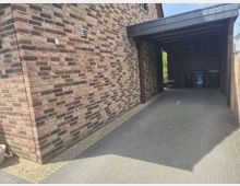 Überdachte Einfahrt neben einem Backsteinhaus mit gepflastertem Boden, Holzbalken und einem flachen, schindelgedeckten Dach. Am Ende des Carports stehen mehrere Mülltonnen, rechts begrenzt von einem Holzzaun.