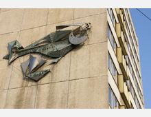 Relief aus Metall in Form eines stilisierten Fisches an der Fassade eines sandfarbenen Plattenbaus mit mehreren Fenstern. Der klare Himmel bildet den Hintergrund.