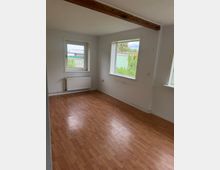 Leerer Wohnraum mit hellem Laminatboden, weißen Wänden und einem sichtbaren Holzbalken an der Decke. Zwei Fenster mit Blick ins Grüne sowie ein Heizkörper unter einem Fenster prägen die Raumseite.