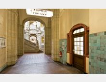 Eingangsbereich eines historischen Gebäudes mit hohen, sandfarbenen Steinsäulen und einer dekorativen Treppe, die zu kunstvollen Bögen mit Stuckverzierungen führt. Auf der rechten Seite befindet sich eine Holztür mit gläsernen Sprossenfenstern, umgeben von türkisfarbenen Fliesenwänden.