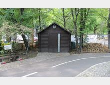 Kleines dunkles Holzhäuschen mit Metalltür in einem von Bäumen umgebenen Bereich. Im Vordergrund verläuft ein gepflasterter Weg und eine asphaltierte Straße mit auffälliger Markierung; links neben dem Häuschen befindet sich ein Bushaltestellenschild.