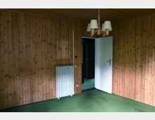 Ein leerer Raum mit holzvertäfelten Wänden und Decke, grünem Teppichboden, einem weißen Heizkörper an der Wand und einer geöffneten weißen Tür, die in einen dunkleren Bereich führt. Eine schlichte Hängelampe mit vier Lampenschirmen hängt von der Decke.
