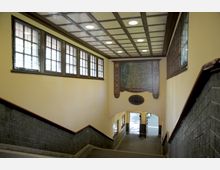 Treppenabgang in einem historischen U-Bahnhof mit gelben Wänden, einer Holzdecke im Kassettendesign und einem großen Wandbild mit einer Karte und der Beschriftung "Dahlem". Die Fenster entlang der Wand bestehen aus kleinen gläsernen Feldern, und am unteren Ende des Abgangs führen Türen ins Freie.