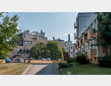 Wohnsiedlung mit gelb-weißen Balkonen auf der rechten Seite, im Hintergrund ein graues, modernistisches Industriegebäude mit zahlreichen Schornsteinen. Dazwischen sind grüne Bäume, parkende Autos und eine gepflasterte Auffahrt zu erkennen.