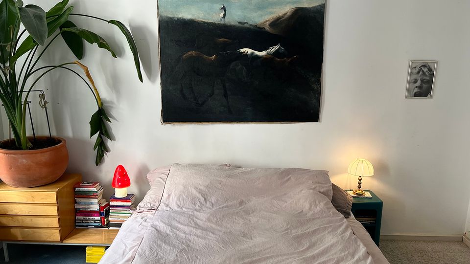 Minimalist bedroom with a low bed and light gray bedding against a white wall, featuring a large dark landscape painting above the headboard. A large potted plant sits on a wooden cabinet beside stacks of books and a small red lamp, with a bedside table holding a lit lamp on the other side.