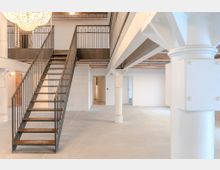 Offener, loftartiger Innenraum mit weiß gestrichenen Wänden und einer Metalltreppe, die zu einer Galerieebene führt. Sichtbare Ziegeldecke, weiße Säulen mit Bolzen und ein großer Kronleuchter sind zentrale architektonische Merkmale.