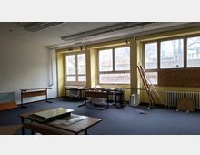 Leerer, teilweise ausgeräumter Klassen- oder Büroraum mit großen Fensterfronten, Heizkörpern darunter und sichtbaren Deckenleuchten. Auf dem Teppichboden stehen einzelne Tische, gestapelte Möbelteile und Holzplatten; draußen sind kahle Bäume durch die Fenster erkennbar.