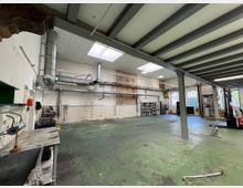 Eine große, leere Werkstatthalle mit grünen, abgenutzten Bodenflächen und sichtbaren Metallträgern an der Decke. An der Wand befinden sich Rohre und Lüftungsleitungen, links steht eine Werkbank, und im Hintergrund sind Regale mit Materialien sowie große Fenster und Tore sichtbar.