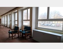 Langer Innenraum mit einer Reihe großer Fenster entlang der Wand, braunem Boden und spärlicher Möblierung mit zwei Sesseln und einem Tisch. Durch die Fenster sind eine Straße mit geparkten Autos sowie gegenüberliegende Gebäude und kahle Bäume zu sehen.