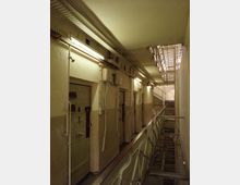 Ein Innenraum eines Gefängnisses mit einem schmalen Gang, flankiert von Zellentüren auf der linken Seite und einem Geländer auf der rechten Seite. Oberhalb des Geländers sind Gitter und Lampen sichtbar, die den schlichten, funktionalen Charakter des Gebäudes betonen.