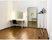 Ein Umkleideraum mit Holzboden, einem weißen Schminktisch mit beleuchtetem Spiegel und einem schwarzen Stuhl. Rechts im Bild befindet sich ein Kleiderständer auf Rollen, daneben ein großer Wandspiegel, der einen angrenzenden Raum reflektiert.