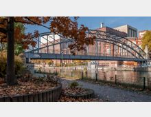 Eine Metallbrücke mit dekorativem Geländer erstreckt sich über einen Fluss, umgeben von Backsteingebäuden und herbstlich gefärbten Bäumen. Im Vordergrund liegt ein Weg mit Laub bedeckt, eingerahmt von Grünflächen und einem Baum.