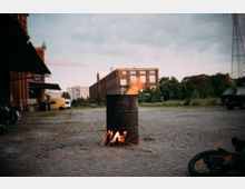 Ein offener Platz mit Kopfsteinpflaster, auf dem in einem Metallfass ein Feuer brennt. Im Hintergrund sind eine rote Backsteinfabrik, moderne Wohngebäude, ein gelber Lieferwagen sowie Bäume und Strommasten zu sehen.