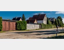 Straßenszene in einem Wohngebiet mit Einfamilienhäusern, niedrigen Hecken und Zäunen; im Vordergrund eine unbefestigte Straße mit Pollern, im Hintergrund mehrere Häuser mit roten und schwarzen Dächern.