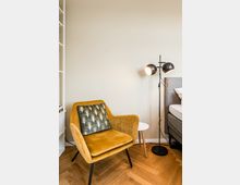 Gemütliche Zimmerecke mit einem senfgelben Sessel und gemustertem Kissen auf Parkettboden, daneben ein kleiner runder Beistelltisch und eine schwarze Stehlampe mit zwei kugelförmigen Leuchten. Am Rand sind ein weißes Regal und ein Teil eines Betts mit grauem Kopfteil zu sehen.