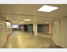 Unterführung mit weiß gekachelten Wänden, hell beleuchteter Decke und gefliestem Boden, links ein Treppenaufgang, vorne geschlossene Metallrollläden und eine rote Tür.