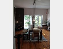 Helles Esszimmer mit Holzboden, einem großen Holztisch und Stühlen mit Fellauflagen. Im Hintergrund gibt es bodentiefe Fenster mit Blick auf einen Garten, und eine moderne Hängelampe hängt über dem Tisch.