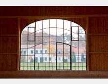 Großes, bogenförmiges Fenster mit Metallstreben in einem Holzgebäude, mit Blick auf eine Grünfläche, weiße Häuser mit roten Dächern und eine Baumreihe im Hintergrund.