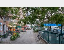 Straßenszene mit einem Gehweg, flankiert von Geschäften, Cafés, geparkten Fahrrädern und Bänken. Im Vordergrund befindet sich das gelb-blaue Schild der U-Bahn-Station „U-Bhf Amrumer Straße“, umgeben von Bäumen und einem Geländer.