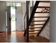 Flur mit geöffneter weißer Haustür, die nach außen führt, einem Terrakotta-Fliesenboden und einer modernen Treppe aus Holzstufen mit schwarzem Metallgeländer. Im Vordergrund befinden sich Kleiderbügel an einer Halterung unter der Treppe.