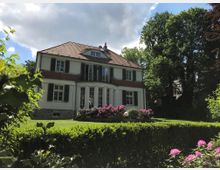Zweistöckiges Haus im Landhausstil mit heller Fassade, roten Dachziegeln und grünen Fensterläden, umgeben von einem gepflegten Garten mit blühenden Sträuchern und Hecken; im Hintergrund Bäume und ein teilweise bewölkter Himmel.