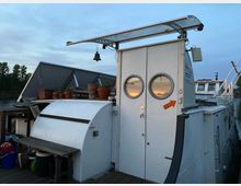 Ein Boot mit weißem Aufbau, auf dem sich zwei runde Fenster in geschlossenen Türen befinden. Auf dem Dach stehen Blumentöpfe, und im Hintergrund sind eine Schiffsleiter sowie ein Fluss mit Bäumen am Ufer sichtbar.